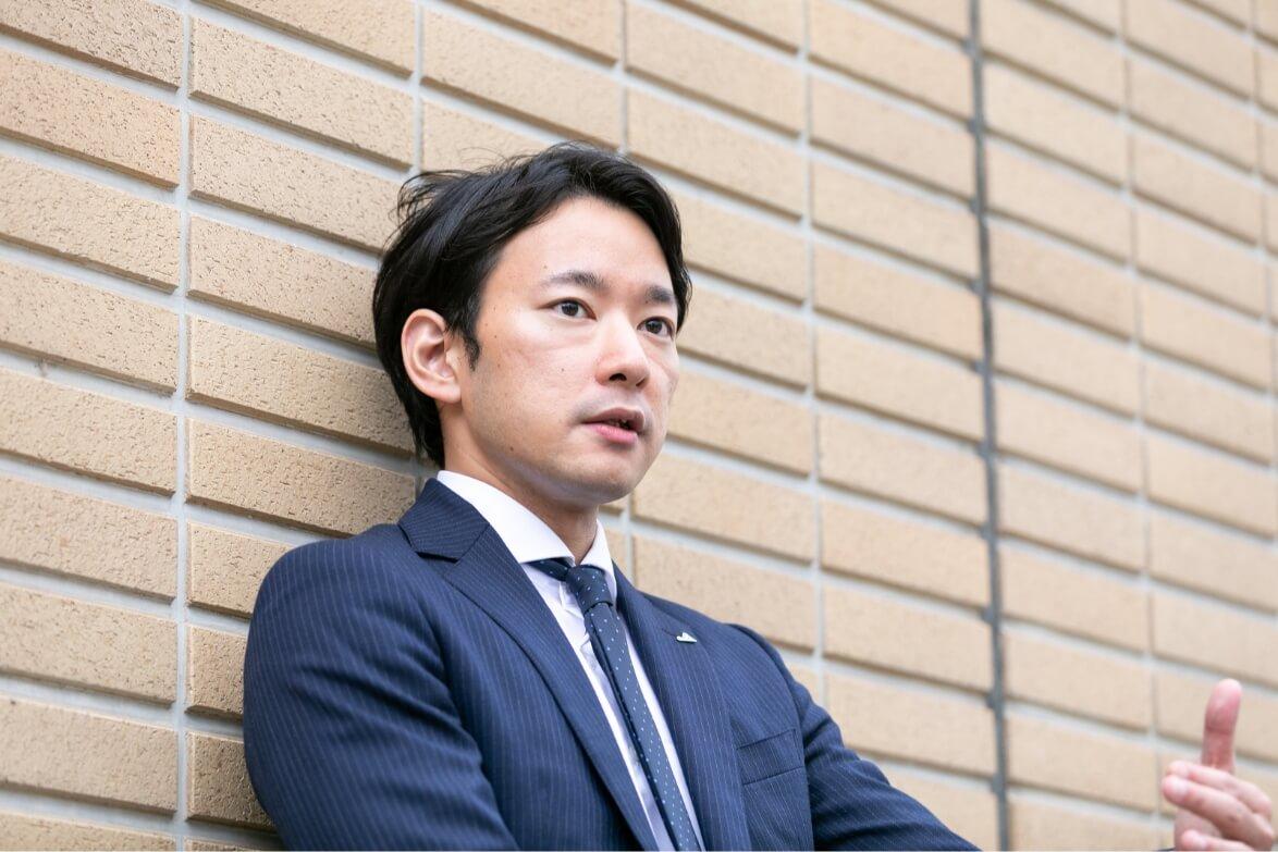 屋外のレンガ壁を背後にインタビューに答える長野県信連法人融資の男性職員
