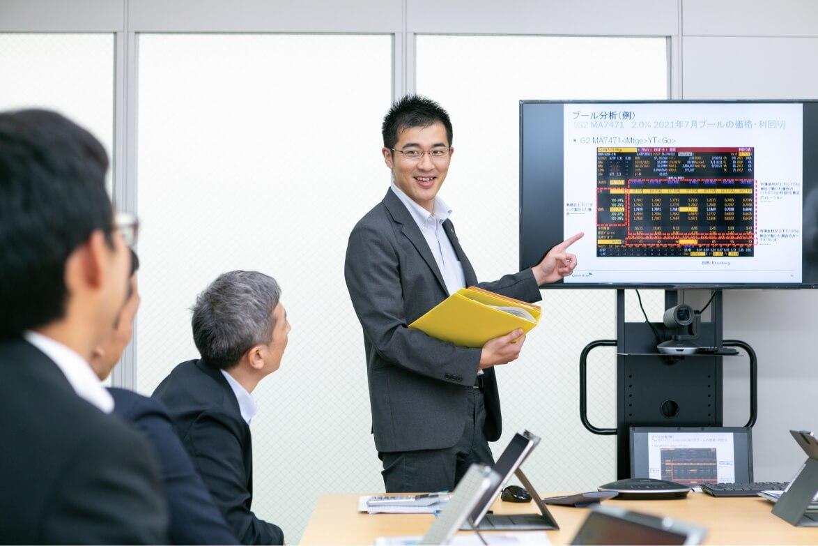 立て式モニターの前で画面に指をさし、チームに最新情報を報告する長野県信連証券運用の男性職員