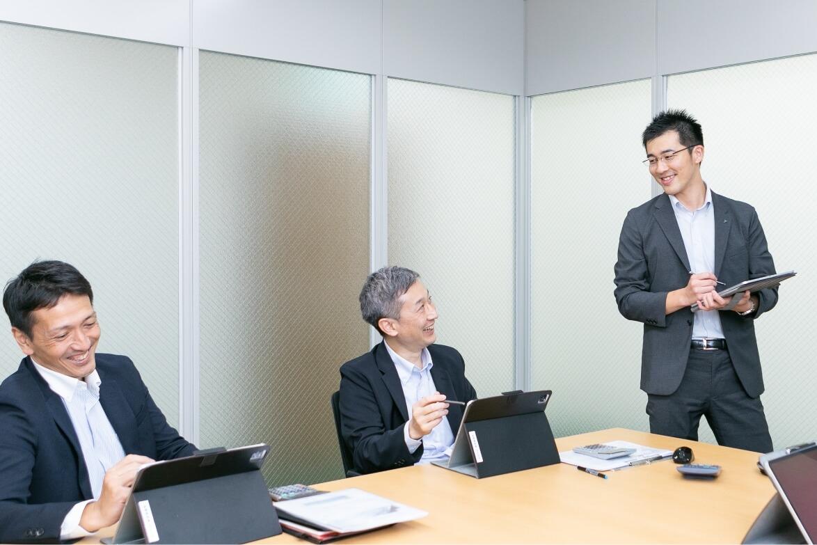 I-padをチェックしながらテーブルに座る上司2名に立ちながら話しかける長野県信連証券運用の男性職員