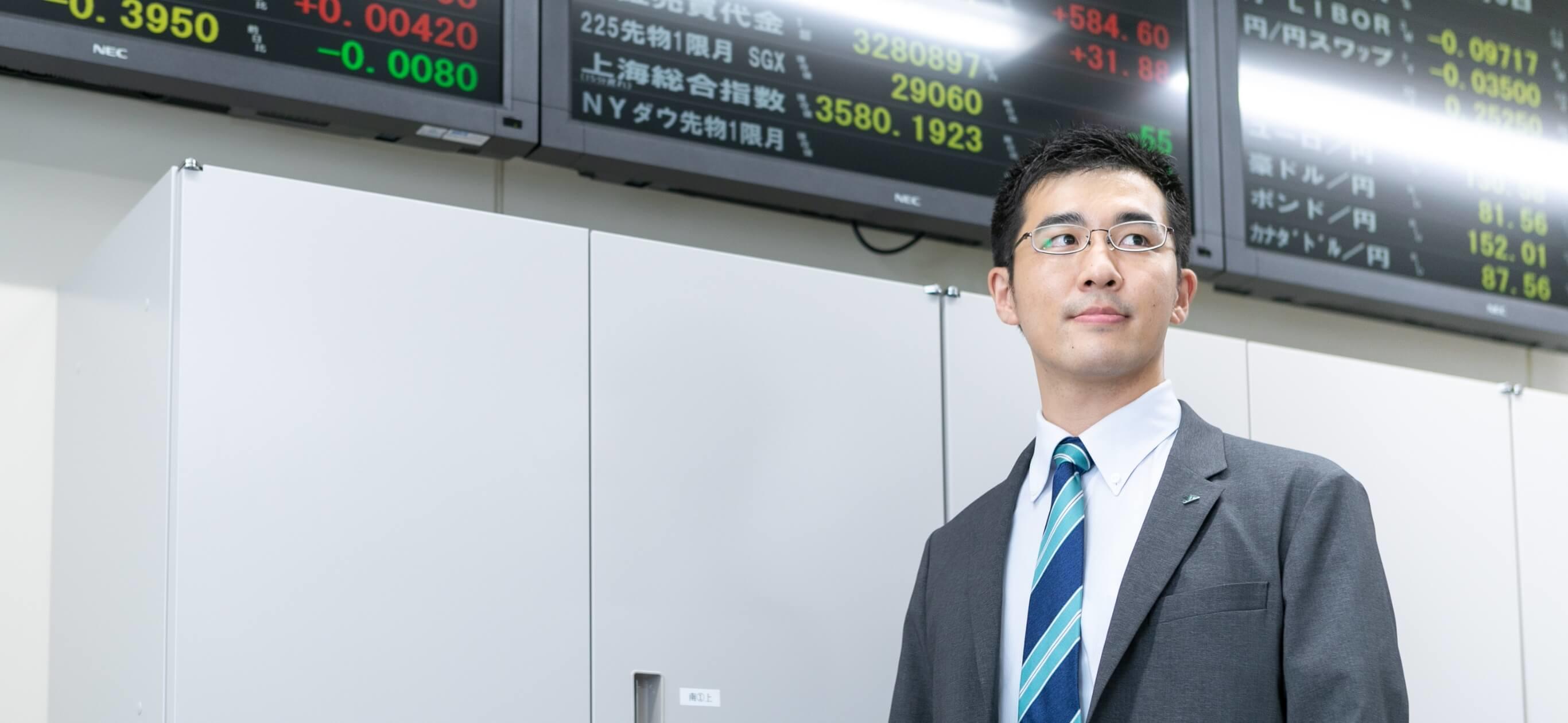 為替レート電光掲示板を背景に立つ長野県信連証券運用の男性職員