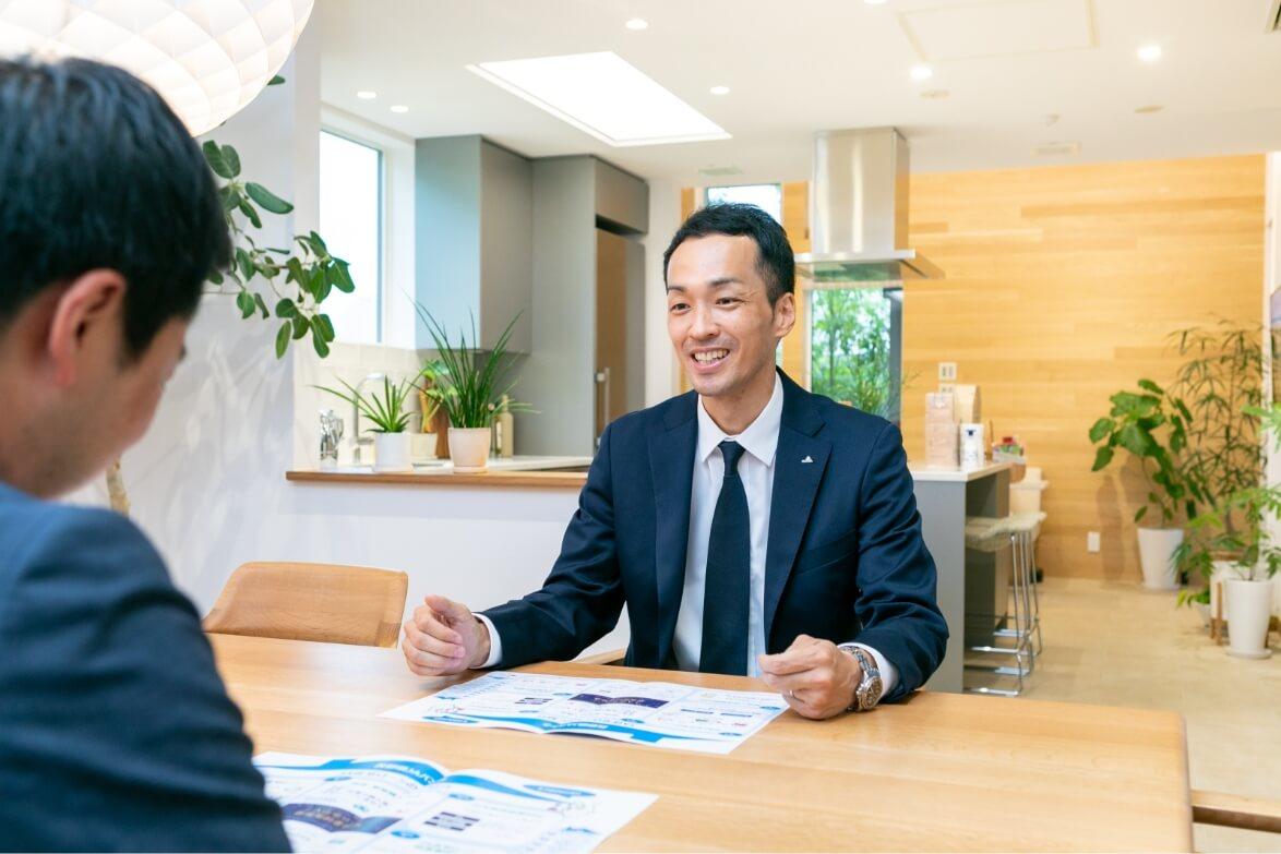 住宅展示場のダイニングテーブルに座り、担当者と商談をする長野県信連ローン推進の男性職員