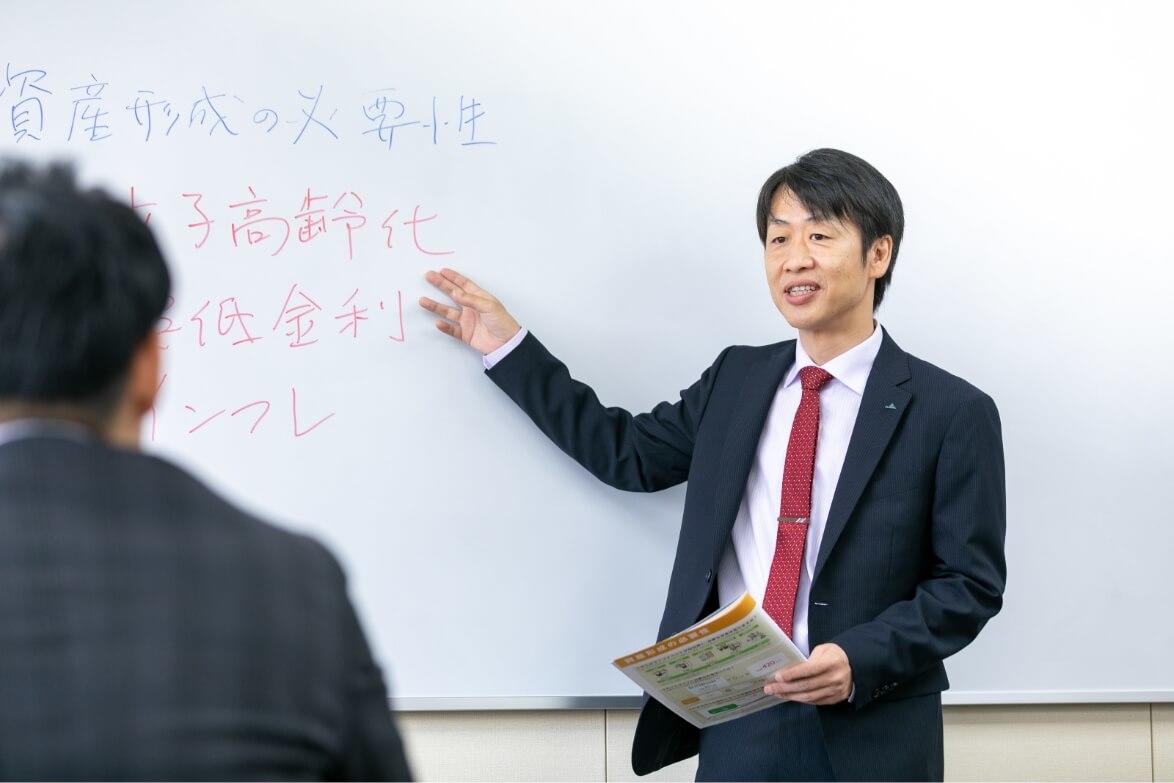 研修中に白板に書いた文字を指差し、座っている職員に話しかける長野県信連ライフサポート推進の男性職員