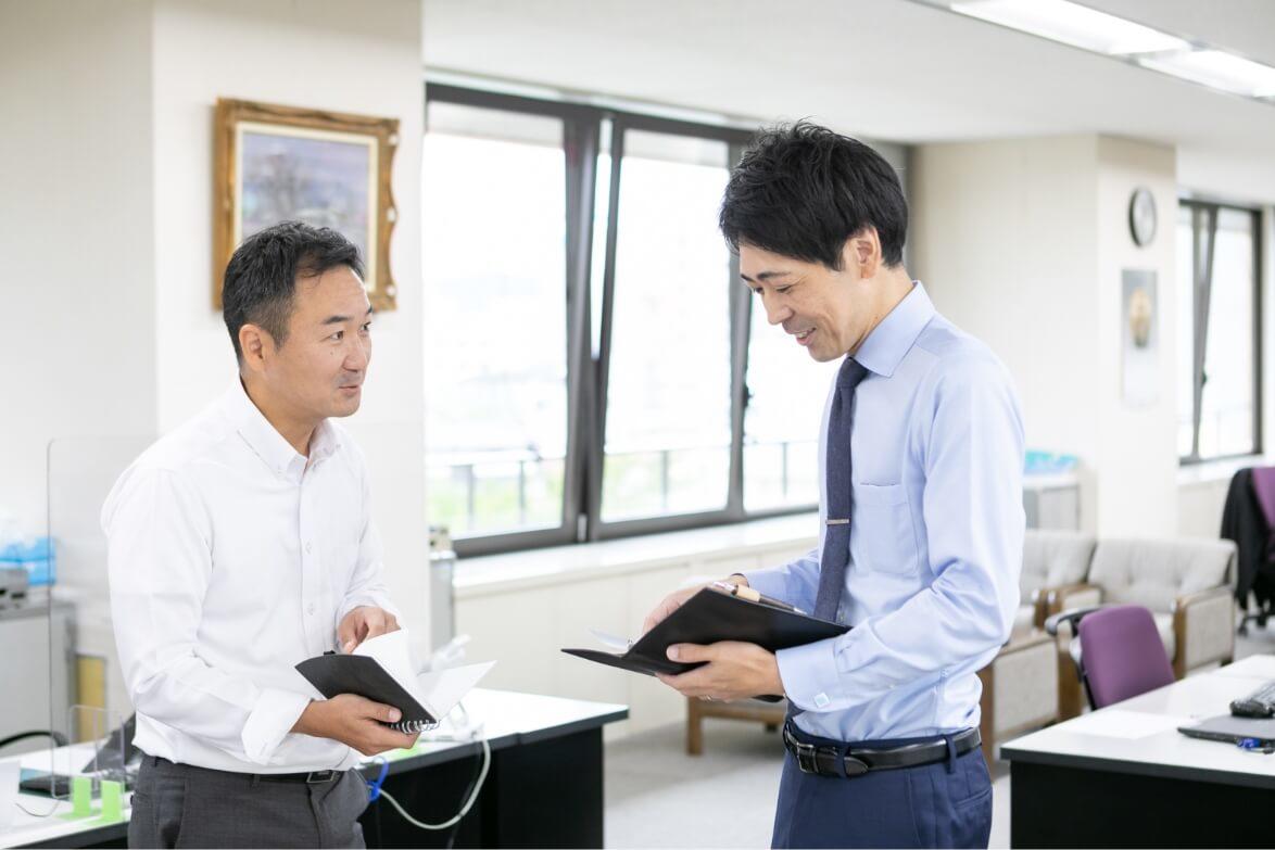 オフィスで立ちながらノートを広げ、上司と談笑をしている長野県信連アグリビジネス推進の男性職員