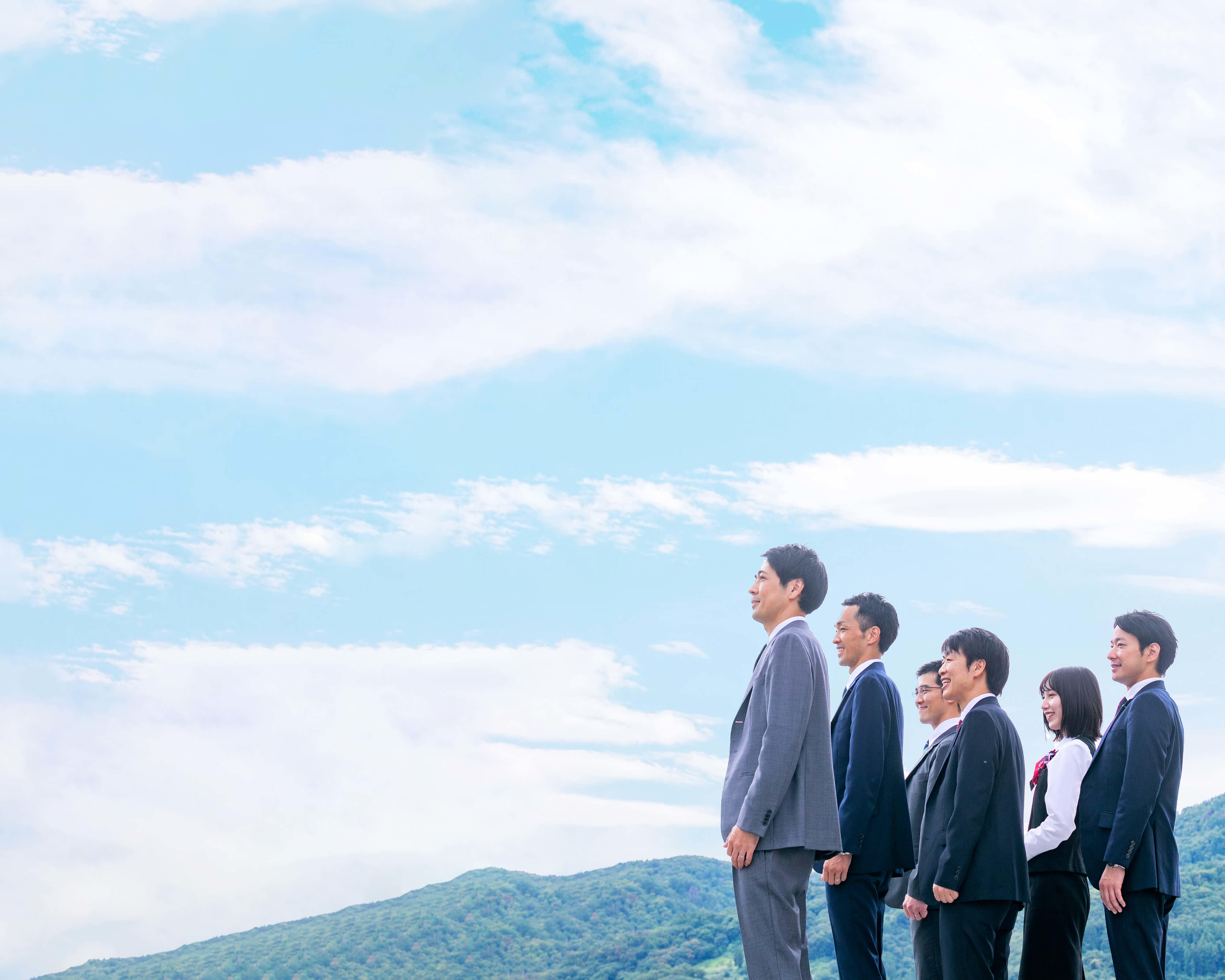 青空と山並みを背景に笑顔で長野市街地を望む長野県信連職員6名