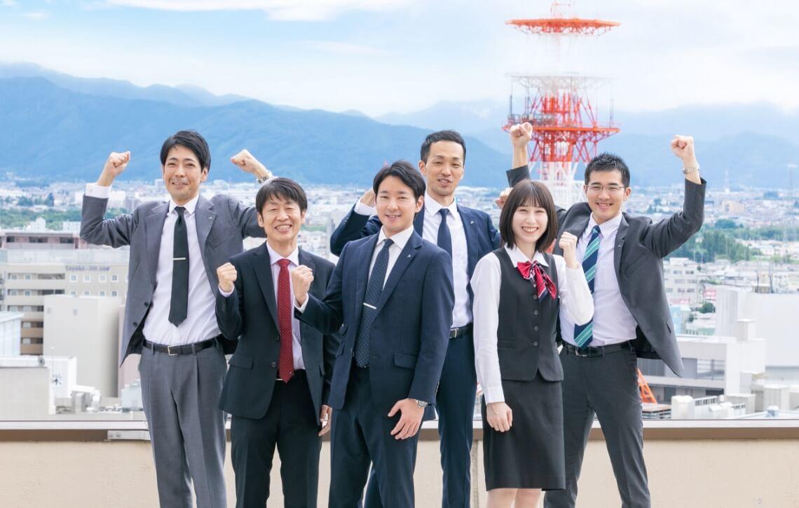 長野市街地のビル群と青空を背景に、ガッツポーズをして立つ長野県信連職員6名
